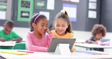 Okulda, iki kız tablet kullanıyor ve sınıfta gülümsüyor, öğrenmenin tadını çıkarıyorlar. Eğitim, teknoloji, öğrenim, çocuklar, arkadaşlık, dijital, değiştirilmemiş, yavaş çekim