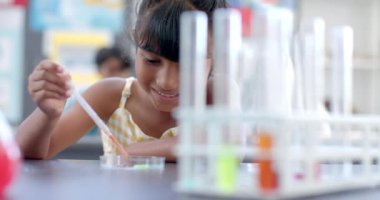 Okuldaki bir sınıf ortamında, melez genç bir kız bir bilim deneyine odaklanır. Koyu renk saçları var, sarı bir elbise giyiyor ve dikkatle bir damlatıcı kullanıyor, değiştirilmemiş, yavaş çekim..