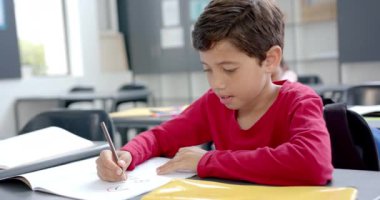 Okulda, kırmızı tişörtlü bir çocuk sınıftaki bir deftere yazı yazıyordu. Sarı bir masada otururken, ödevini bitirmeye odaklanmış görünüyor, değiştirilmemiş, ağır çekimde..
