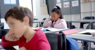 Okulda, çocuklar sınıftaki işlerine odaklanmış durumdalar. Bir kız, derin düşüncelere dalmış, kafası elinde, rengarenk resimler ve matematik işaretleriyle çevrili, değişmemiş, yavaş çekim.