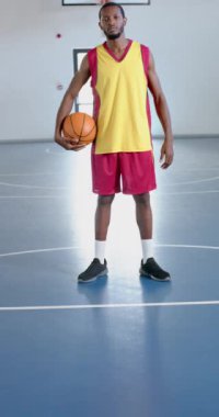 Dikey video: Basketbolu tutan, kapalı sahada spor üniformalı bir erkek atlet. kapalı alan, basketbol sahası, yarışma, atletik, fitness, antrenman