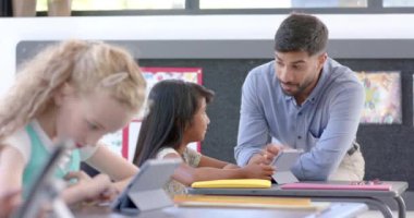 Genç Asyalı bir adam, iki ırklı bir kıza okuldaki çalışmalarında tabletle yardımcı oluyor. Modern eğitim ortamını gösteren bir tablete odaklanırlar, ağır çekimde..