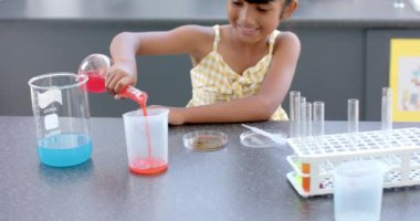 Bir okul laboratuvarında, melez bir kız bir kimya deneyi yapıyor. Etrafı deney tüpleri ve deney tüpleriyle çevrili kırmızı bir sıvı dolduruyor. Koyu renk saçı bir yayla bağlanmış, değiştirilmemiş.