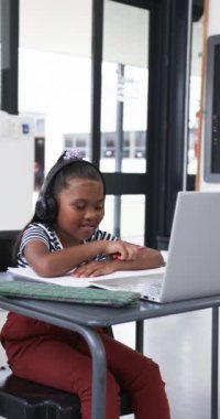 Dikey video: Bir okul ortamında, genç bir Afrikalı Amerikalı öğrenci elini kaldırıyor. Koyu renk saçları var, kulaklık takıyor ve masasındaki dizüstü bilgisayarla etkileşime geçiyor.