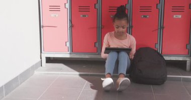 Dolapların yanında oturan Afro-Amerikan kız tablet kullanıyor ve okul koridorunda gülümsüyor. Eğitim, teknoloji, öğrenci, öğrenme, gençlik, ergenlik