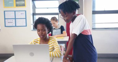 Okulda, iki kız öğrenci laptopta iş birliği yaparken başka bir öğrenci yanımızdan geçiyor. Eğitim, öğrenim, işbirliği, teknoloji, takım çalışması, öğrenme