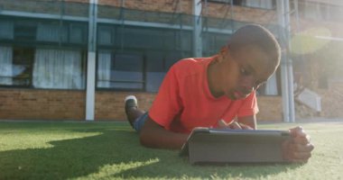 Çimlerin üzerinde uzanan, okul binasının dışında okul ödevleri için tablet kullanan bir çocuk. Eğitim, teknoloji, açık hava, öğrenim, çocukluk, öğrenim