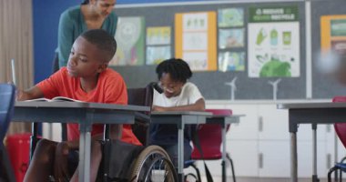 Okulda, tekerlekli sandalyedeki çocuk, öğretmen asistanı ile deftere yazı yazıyor. Eğitim, öğrenim, sınıf, kapsama, öğretim, engellilik