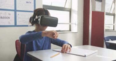 Okulda, masada defter yazarken VR kulaklık kullanan bir çocuk. Eğitim, teknoloji, sanal gerçeklik, öğrenme, sınıf, yenilik