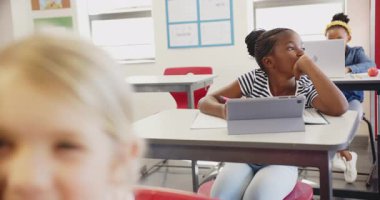 Okulda, masada tabletle oturan bir kız, sınıfta düşünceli görünüyor. Eğitim, öğrenim, öğrenim, teknoloji, çocukluk