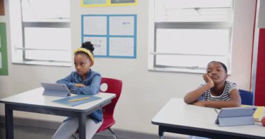 Okulda, masada tablet kullanan bir kız ve yanında esneyen başka bir kız. Eğitim, sınıf, teknoloji, öğrenme, çocukluk, öğrenciler