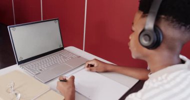 Okulda, kulaklık takan Afro-Amerikalı genç çocuk dizüstü bilgisayar ve not defteriyle ders çalışıyor, fotokopi çekiyor. Eğitim, teknoloji, öğrenme, konsantrasyon, çocukluk, online