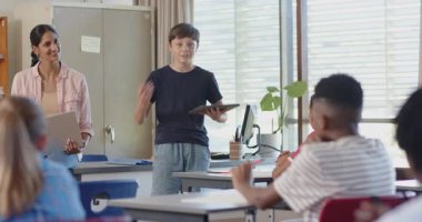 Okulda, elinde tablet olan bir çocuk sınıf arkadaşlarına gülümseyen bir öğretmen hediye ediyordu. Eğitim, sınıf, öğrenim, sunum, öğrenciler, teknoloji