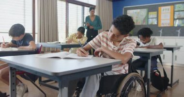 Okulda, tekerlekli sandalyedeki çocuk deftere yazı yazıyor, öğretmen öğrencilere yardım ediyor. Eğitim, kapsama, engellilik, öğrenme, sınıf, akıl hocalığı