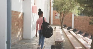 Bankta oturan, sırt çantalı Afrikalı Amerikalı kız okul binasının önünde bekliyor. Öğrenci, eğitim, açık hava, çocukluk, okula dönüş.