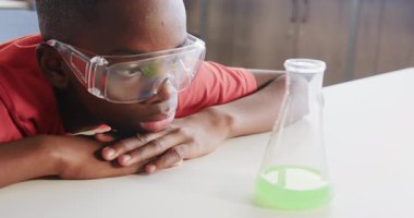 Okulda, koruyucu gözlük takan bir çocuk beher içinde yeşil sıvı gözlüyor. Bilim, deney, eğitim, sınıf, öğrenme, kimya