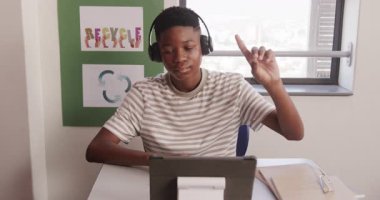 Okulda, Afrika kökenli Amerikalı genç bir çocuk kulaklık takıyordu. Masa başında çalışmak için tablet kullanıyordu. Eğitim, teknoloji, öğrenme, konsantrasyon, öğrenci, sınıf