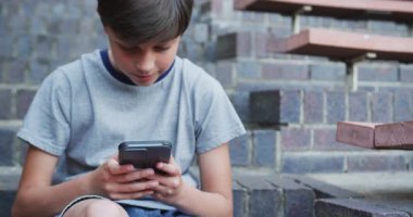 Okul merdivenlerinde oturmuş, tatilin tadını çıkarırken akıllı telefon kullanan gülümseyen çocuk. Gençlik, teknoloji, rahatlama, dijital, bağlantı, öğrenci