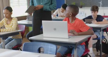 Öğretmen sınıfta dizüstü bilgisayarla öğrenciye yardım ediyor, diğer öğrenciler okuyor. Eğitim, öğrenim, öğretim, teknoloji, destek, akademik