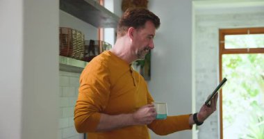 Video görüşmesi sırasında elinde tablet ve kahve fincanı tutan gülümseyen adam. Teknoloji, ev ofisi, sanal, iletişim, online toplantı, dijital