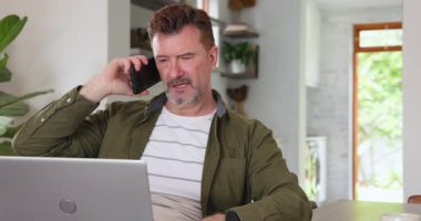 Evde çalışan, dizüstü bilgisayar kullanan ve akıllı telefondan konuşan kıdemli bir adam. Ev ofisi, teknoloji, uzaktan çalışma, kıdemli yetişkin, telekomünikasyon, iletişim.