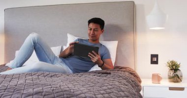 Evde dinlenen Asyalı adam, rahat yatakta tabletin tadını çıkarıyor. Teknoloji, rahatlama, rahatlama, dijital, yaşam tarzı, konfor