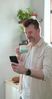 Dikey video: Yaşlı adam evde akıllı telefondan kitap okurken kahvenin tadını çıkarıyor. Teknoloji, yaşam tarzı, emeklilik, rahatlama, dijital, boş zaman