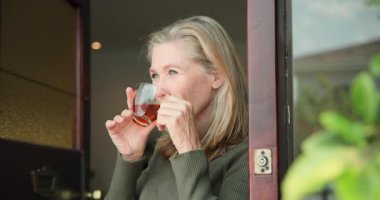 Gülümseyen son sınıf öğrencisi kadın pencerenin önünde çaydan zevk alıyor, evde huzurlu bir anı kucaklıyor. Rahatlama, sükunet, yaşlılık, yaşam tarzı, içecek, kapalı alan