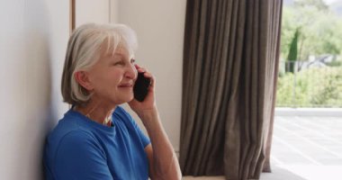 Mavi gömlekli yaşlı kadın evde akıllı telefondan konuşuyor, düşünceli görünüyor. Yaşlılar, iletişim, teknoloji, yaşam tarzı, yerli, düşünceli