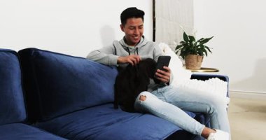 Gülümseyen Asyalı adam akıllı telefonda mavi koltukta köpekle sohbet ediyor. Teknoloji, mutluluk, evcil hayvan, iletişim, yaşam tarzı, konfor