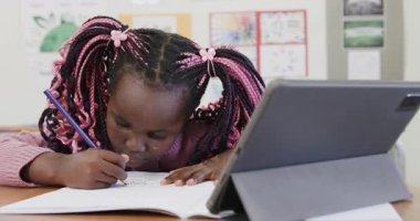 Sınıfta, not defterine yazı yazan genç bir kız, tabletle okul ödevlerine odaklanmış. Eğitim, öğrenim, teknoloji, dijital öğrenme, kadın kız çocuğu, konsantrasyon