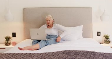 Üst düzey kadın dizüstü bilgisayar kullanarak yatakta dinleniyor, evde huzurlu vakit geçiriyor. Yaşlı, teknoloji, rahatlama, rahatlık, konfor, yaşam tarzı