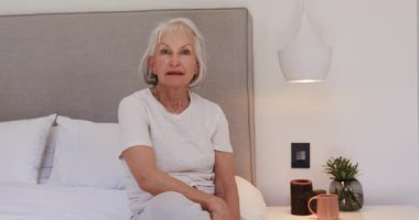 Evde yatakta oturmuş, huzurlu bir sabah geçirirken kıdemli bir kadın gülümsüyor. Yaşlılık, rahatlama, yaşam tarzı, huzur, mutluluk.