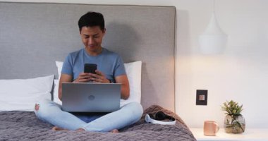 Evdeki Asyalı adam dizüstü bilgisayar, kulaklık ve akıllı telefon kullanarak yatakta oturuyor. Teknoloji, uzaktan çalışma, çoklu görev, dijital, iletişim, gelişigüzel