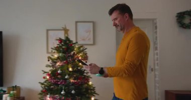 Oturma odasında akıllı telefon kullanan, televizyon ve çiçeklerin yanında gülümseyen kıdemli bir adam. teknoloji, ev, rahatlama, rahatlama, yaşam tarzı, iç mekan