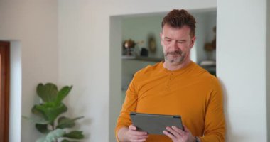 Yaşlı adam evde tablet kullanıyor, ekrana odaklanıyor, turuncu gömlek giyiyor. Teknoloji, yaşlılar, yaşam tarzı, gündelik, konsantrasyon, dijital