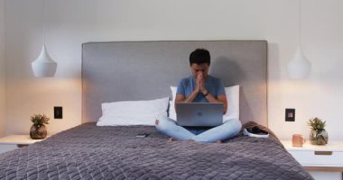 Asyalı adam evde yatakta otururken akıllı telefon ve dizüstü bilgisayar kullanıyor. Teknoloji, dijital, çoklu görev, yaşam tarzı, iletişim, aygıt