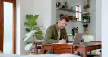 Evde çalışan kıdemli bir adam, not yazıyor ve mutfakta dizüstü bilgisayar kullanıyor. Uzaktan çalışma, telekomünikasyon, teknoloji, olgun yetişkin, ev ofisi, verimlilik