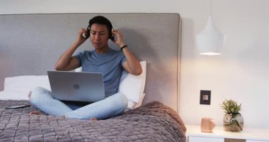 Evde dinlenirken, Asyalı adam akıllı telefon ve dizüstü bilgisayar kullanıyor, müzikten zevk alıyor. Teknoloji, eğlence, yaşam tarzı, rahatlama, kulaklık, dijital