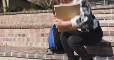 Merdivenlerde oturan liseli kız yanında mavi bir sırt çantasıyla kitap okuyor. Eğitim, merdiven, genç kız, bilgi, boş zaman, çocukluk