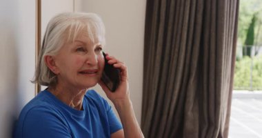 Mavi gömlekli yaşlı kadın akıllı telefonuyla konuşuyor, evde gülümsüyor. Telekomünikasyon, iletişim, teknoloji, yaşam tarzı, gelişigüzel, mutluluk