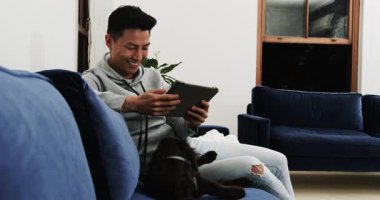 Evde dinlenirken, Asyalı bir adam kucağında köpeğiyle tablet kullanıyor, sıcak bir şekilde gülümsüyor. Teknoloji, dijital cihaz, evcil hayvan, eğlence, oturma odası, arkadaşlık.