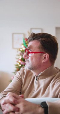 Dikey video: Gülümseyen, gözlüklü yaşlı adam evde Noel ağacının yanında oturuyor. Tatil, neşeli, kutlama, gelenek, kış, neşe