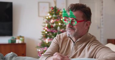 Süslü Noel ağacının yanında oturan, neşeli gözlük takan gülümseyen son sınıf öğrencisi. Tatil, kutlama, yaşlı, neşeli, gelenek, mutluluk