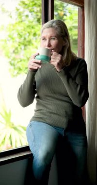 Dikey video: Gülümseyen son sınıf öğrencisi kadın evde kahvesini yudumluyor, rahatlıyor. Rahatlama, sabah, rahatlık, rahatlık, yaşam tarzı, olgun bir kadın.