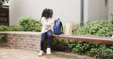 Genç bir kız bankta oturuyor, okulun yanındaki sırt çantasını açıyor, moladan zevk alıyor, fotokopi çekiyor. Öğrenci kız, eğitim, mola, kız, açık hava, dinlenme