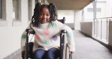 Tekerlekli sandalyedeki gülümseyen kız okul koridorunda eğleniyor, renkli bir kazak giyiyor. Erişilebilirlik, engellilik, kapsayıcılık, bağımsızlık, mutluluk, güçlendirme