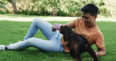 Asyalı bir adam çimenlerde köpek yavrusuyla oynuyor. Parkta güneşli bir günün tadını çıkarıyor. Evcil hayvanlar, arkadaşlık, doğa, eğlence, mutluluk, bağ.