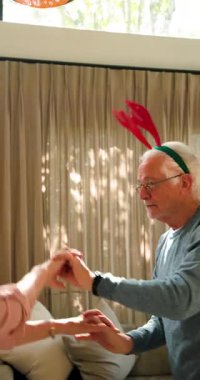 Dikey video: Ren geyiği boynuzu takan yaşlı bir adam Noel boyunca evde neşeyle dans ediyor. Kutlama, festival, tatiller, dekorasyon, kış, gelenekler