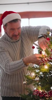 Dikey video: kıdemli adam Noel ağacını evde süslüyor, Noel Baba şapkası takıyor ve gülümsüyor. Tatil, şenlik, kutlama, gelenek, dekorasyon, yaşlılık